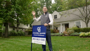 Get Pre-Approved for a Mortgage in Vermont 8 Realtor® John Black standing in front of a residential home, holding a Coldwell Banker Hickok & Boardman yard sign and waving to greet clients