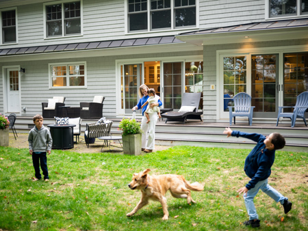 Family and dog playing in a grassy backyard behind a home with a large deck and patio seating, capturing an active outdoor moment at the property.
