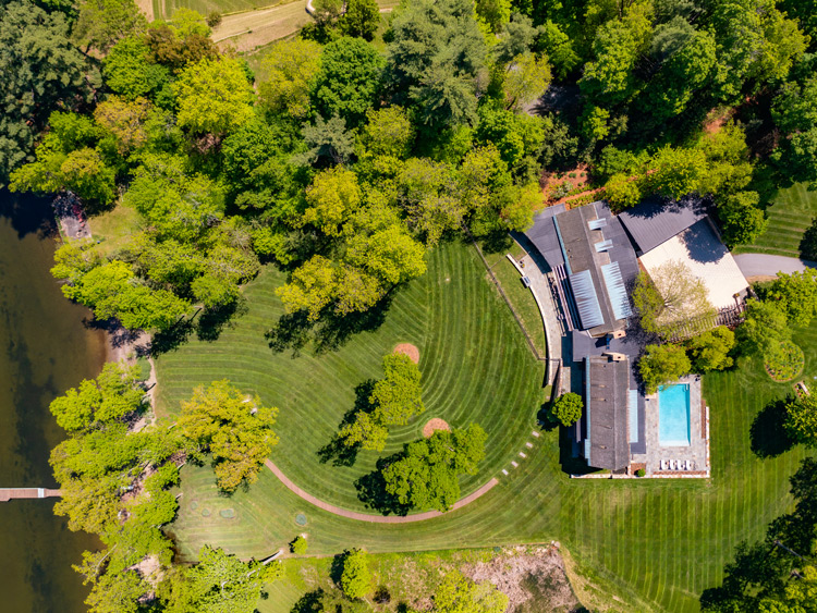 Aerial view of a luxury lakeside property featuring a large home, manicured lawn, outdoor pool, and private shoreline surrounded by mature trees.