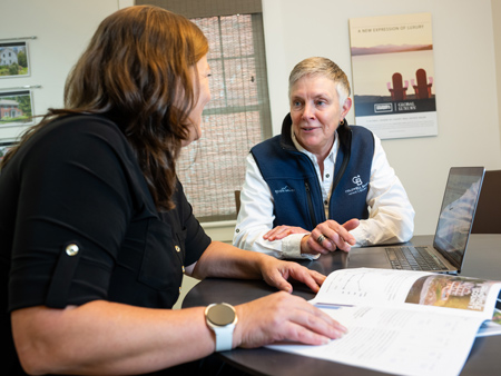 Real estate agent meeting with a homeowner at a table, reviewing a marketing report and property materials on a laptop and printed documents while discussing the marketing performance of the client’s home.