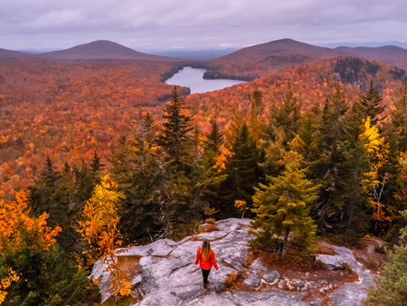 Person standing on a rocky overlook surrounded by vibrant autumn foliage, looking out over a lake and rolling Vermont mountains during peak fall color.